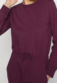 Jumpsuit lungo bordeaux con maniche lunghe, realizzato in tessuto morbido, con scollatura rotonda, vita elastica con cordoncino e polsini aderenti.