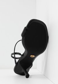 Sandale noire à talons hauts avec des fines lanières, vue du côté de la semelle, positionnée contre des murs blancs au coin.