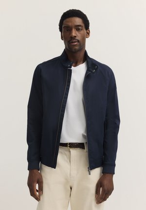 Marineblaue Jacke mit einem Reißverschluss vorne, hohem Kragen und gerippten Bündchen. Getragen über einem weißen Hemd und kombiniert mit hellen Hosen. Glatte Stoffoberfläche.