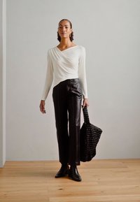 White long-sleeve wrap top, black leather straight-leg pants with stitch detailing, and a woven black bag. Floor with light wood texture.