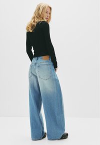 Hellblaue, weit geschnittene Jeans mit einem braunen Lederpatch auf der Rückseite, fünf Taschen und einem verwaschenen Aussehen. Schwarzes Langarmoberteil sichtbar.