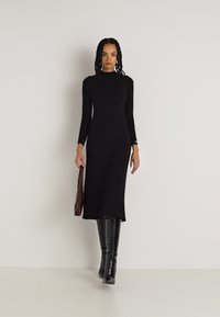 Črna rebrasta pletena obleka z dolgimi rokavi, do kolen, v kombinaciji z visokimi, sijočimi črnimi škornji. Model drži maroon torbico.