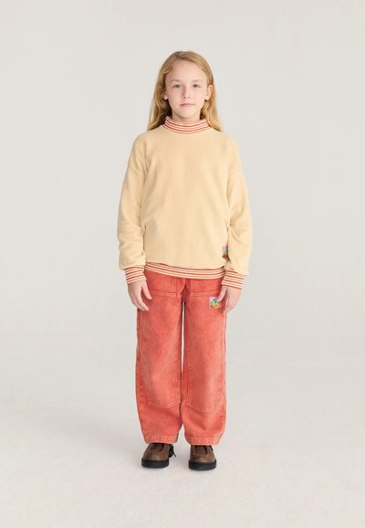 Enfant blond debout, portant un sweat-shirt beige avec col et poignets rayés, pantalon rouge ample et chaussures marron sur fond clair.