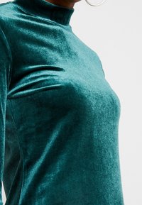 Top de terciopelo verde azulado de cuello alto con una textura suave, que presenta un diseño ajustado y un ligero brillo, resaltando las curvas de la tela.