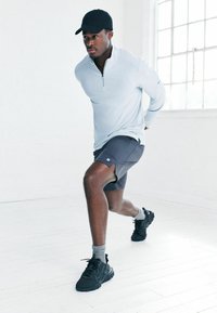 Vīrietis melnā cepurē, gaišā garajās piedurknēs augšējā daļā un tumšās šortos veic priekšējo izsperšanu spilgtā baltā telpā.