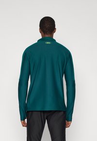 Μπλε-πράσινο αθλητικό πουκάμισο με μακριά μανίκια και ψηλό γιακά, με κίτρινο λογότυπο στο πίσω μέρος και κομψό σχέδιο για εφαρμοστή σιλουέτα.