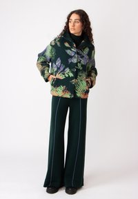 Zelena jakna s cvetnim vzorcem in velikimi gumbi, v kombinaciji z zelenimi širokimi hlačami z modrim obrobjem. Mehka tekstura, sproščen kroj.
