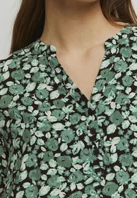 Blusa floreale con sfondo nero, decorata con foglie verdi e fiori. Design con scollo a V, dotata di una patta e tre bottoni neri.