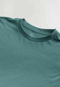 Tricou din bumbac teal, cu guler rotund și mâneci scurte, având o textură netedă și un eticheta cu inscripția "NEXT."