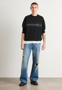 Felpa nera con logo "HUGO" blu, abbinata a jeans strappati blu chiaro e scarpe nere, su uno sfondo neutro.