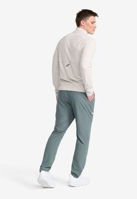 Ljusgrå långärmad tröja med hög krage och logotyp på baksidan, kombinerad med mörkgröna byxor och vita träningsskor. Slät tyg och figurnära design.