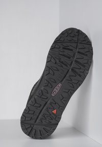 Svart sneakersula med grov mönsterdesign, prydd med en rosa logotyp och röda accenter. Materialet är gummi med en texturerad yta för bättre grepp.