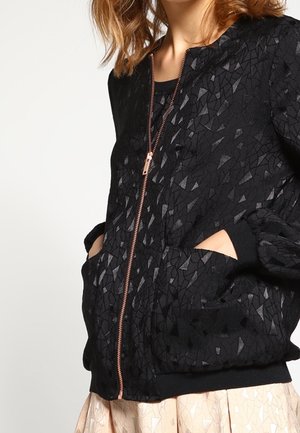 Kvinde iført en sort mønstret jakke med lynlås og hænderne i lommerne, kombineret med et lyst farvet nederdel mod en ensfarvet baggrund.