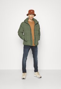 Giacca verde imbottita con zip frontale, T-shirt marrone sotto, jeans scuri e sneaker beige, completata da un cappello a secchiello marrone.