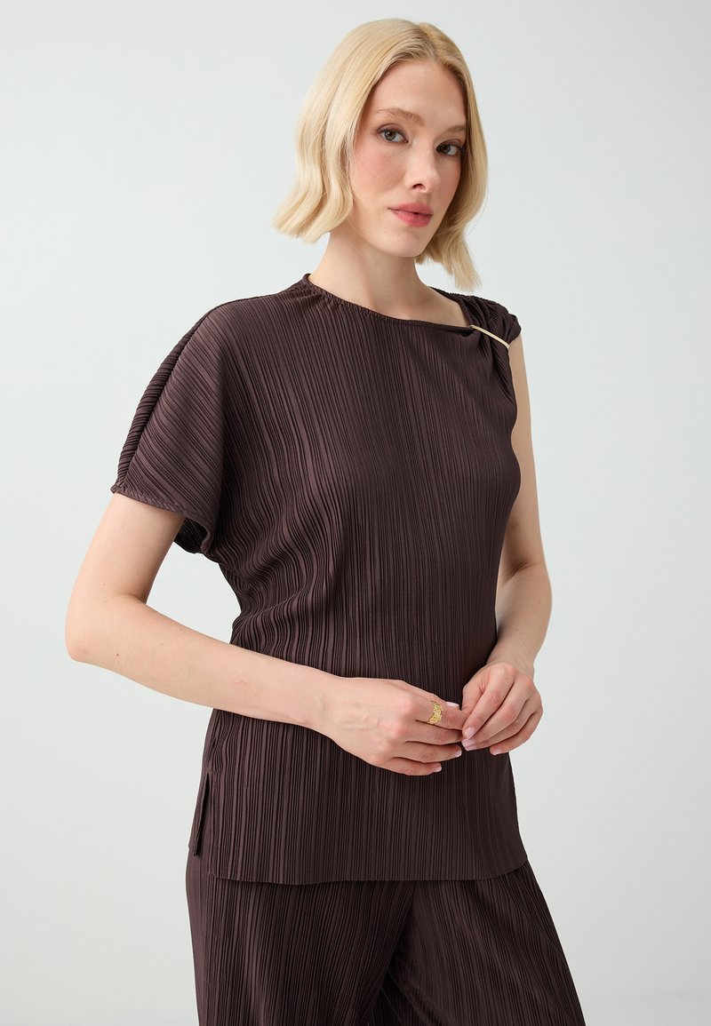 Femme aux cheveux courts et blonds, portant un haut plissé marron foncé et un pantalon assorti, debout devant un fond clair uni.