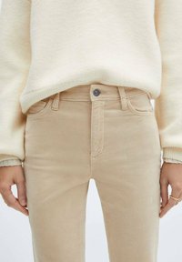 Beige katoenen jeans met een slim fit, voorzien van een metalen knoopsluiting, riemlussen en subtiele stikseldetails. Draag met een crèmekleurige trui.