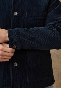 Giacca in velluto a coste blu navy con chiusura a bottoni, piccola tasca sul petto e tessuto a coste testurizzato. Manica con dettaglio a bottone.