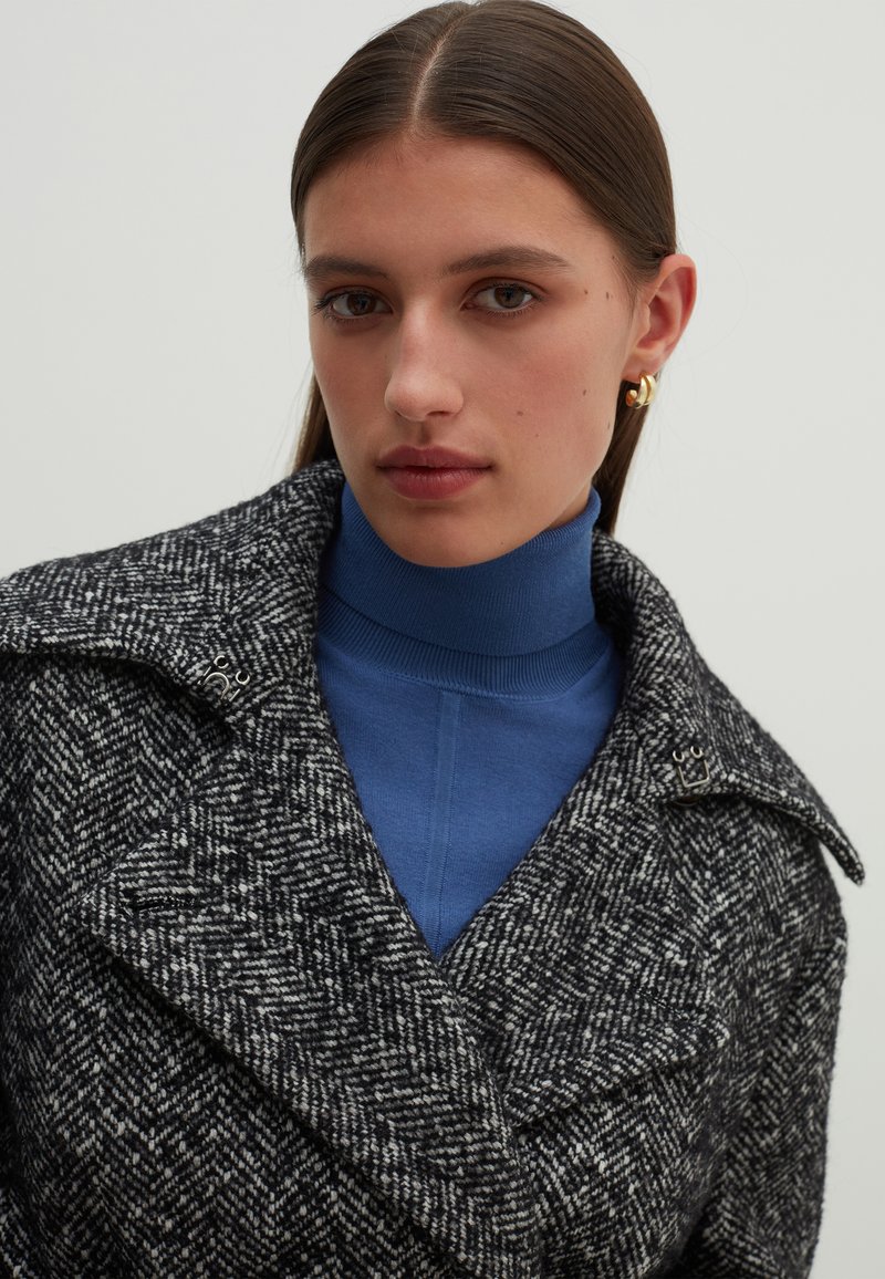 Cappotto in lana bianco e nero a motivo houndstooth con un ampio colletto, hardware argentato, indossato sopra un dolcevita blu. Texture liscia, design strutturato.