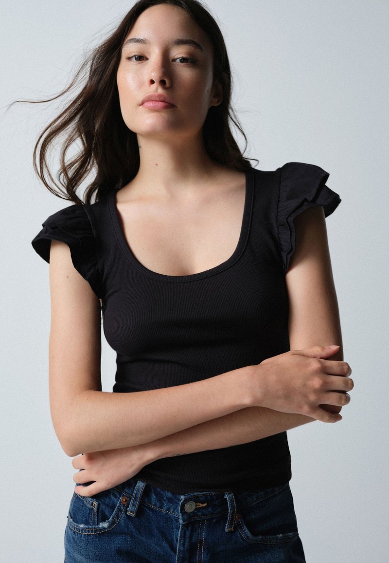 Joven mujer con cabello largo y oscuro, vestida con una blusa negra con mangas abullonadas y vaqueros azules, está de pie con los brazos cruzados.