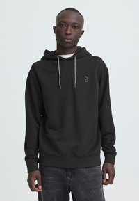 Zwarte hoodie met een getextureerde afwerking, trekkoorden en een logo-accent. Gecombineerd met donkergrijze jeans met slijtage.