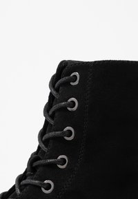 Botín de ante negro con ojales de metal y cordones negros. La vista de cerca enfatiza la textura y los detalles de costura.