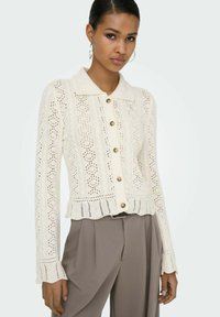 Cardigan crème tricoté avec col, présentant une fermeture à boutons sur le devant, un motif en dentelle et un ourlet festonné. Associé à un pantalon beige taille haute.