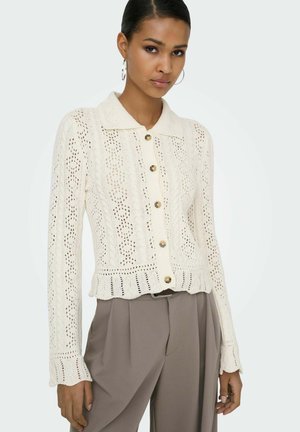 Cardigan crème tricoté avec col, présentant une fermeture à boutons sur le devant, un motif en dentelle et un ourlet festonné. Associé à un pantalon beige taille haute.