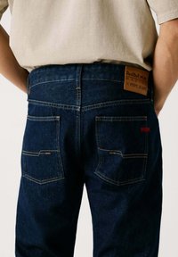 Jeans de mezclilla azul oscuro con una superficie texturizada, que cuentan con dos bolsillos traseros y una etiqueta de marca marrón en la cintura. Costuras amarillas como acento.