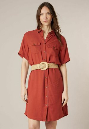 Femme aux longs cheveux bruns portant une robe boutonnée couleur rouille avec des poches poitrine et une large ceinture beige tissée.