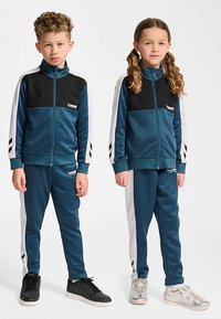 Blå træningsdragter med sorte accenter, som har jakker med lynlåse og bukser med elastik. Sidestriber og logodetalje på brystet.