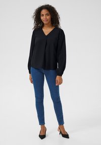 Blouse noire à manches longues avec un col en V et un léger détail plissé, associée à un jean bleu ajusté et des escarpins noirs à bout pointu.