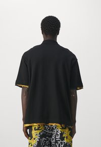 Czarna koszula z krótkim rękawem i kołnierzykiem, z kolorowym wzorem na dole i przy rękawach. Gładka bawełniana tekstura.