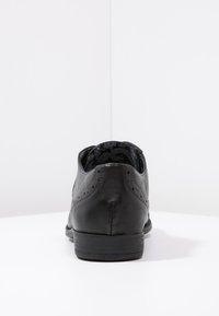 Chaussure habillée en cuir noir avec une forme arrondie, des perforations décoratives et un talon bas texturé. L'intérieur présente une doublure à motif.