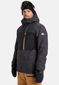 Man in een zwart gespikkelde winterjas met capuchon, zwarte handschoenen, een zwarte beanie, skibril op het hoofd en tan kleurige sneeuwbroek tegen een witte achtergrond.