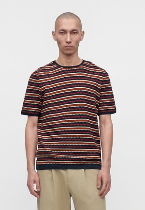 Homme portant un t-shirt à manches courtes col rond avec des rayures horizontales bleu marine, rouge et beige, et un pantalon beige sur fond clair uni.