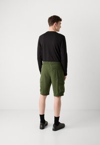 Grønne cargoshorts med flere lommer, elastisk linning, kombinert med en svart langermet topp og svarte sko.
