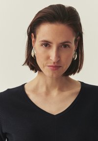 Suéter de punto azul marino con diseño de cuello en V, tejido texturizado y silueta ajustada. Pendientes grandes plateados adornan el cabello corto y oscuro.