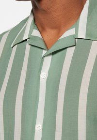 Chemise à manches courtes rayée vert et blanc, dotée d'un col et d'une fermeture à boutons, fabriquée en tissu léger à la texture lisse.