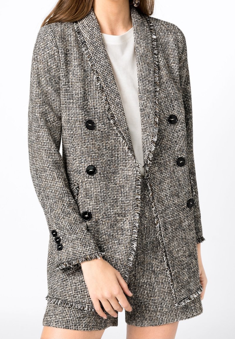Femme portant un blazer croisé à carreaux noirs et blancs avec un short assorti et un haut blanc à col rond.