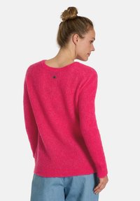 Flauschiger rosa Pullover mit rundem Ausschnitt und gerippter Textur, der eine dezente Knopfdetail hinten aufweist. Kombiniert mit hellblauen Hosen.