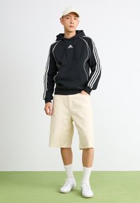 Jovem vestido com uma hoodie preta da Adidas, calções bege, meias brancas, sapatilhas e um boné bege, de pé sobre um chão verde contra uma parede branca.