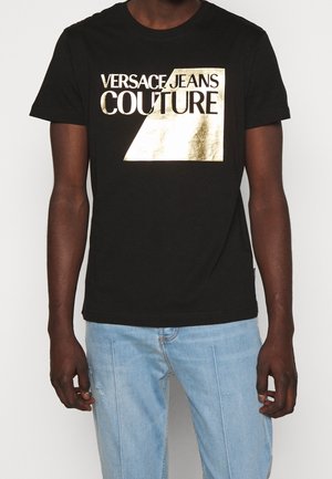 Mężczyzna ubrany w czarną koszulkę z błyszczącym złotym napisem "Versace Jeans Couture" i jasnoniebieskie dżinsy na jednolitym tle.