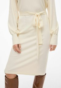 Robe en tricot crème avec des manches longues et une ceinture à nouer à la taille. Présente une finition texturée et une coupe droite à mi-genoux.