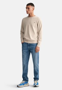 Beige Strickpullover mit Rundhalsausschnitt, kombiniert mit blauen Jeans und blauen Akzenten in den Sneakers. Schlichte und glatte Textur.