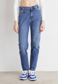 Jeans de mezclilla azul de cintura alta con pierna recta, diseño de cinco bolsillos y costuras en contraste, combinados con zapatillas azul y blancas.