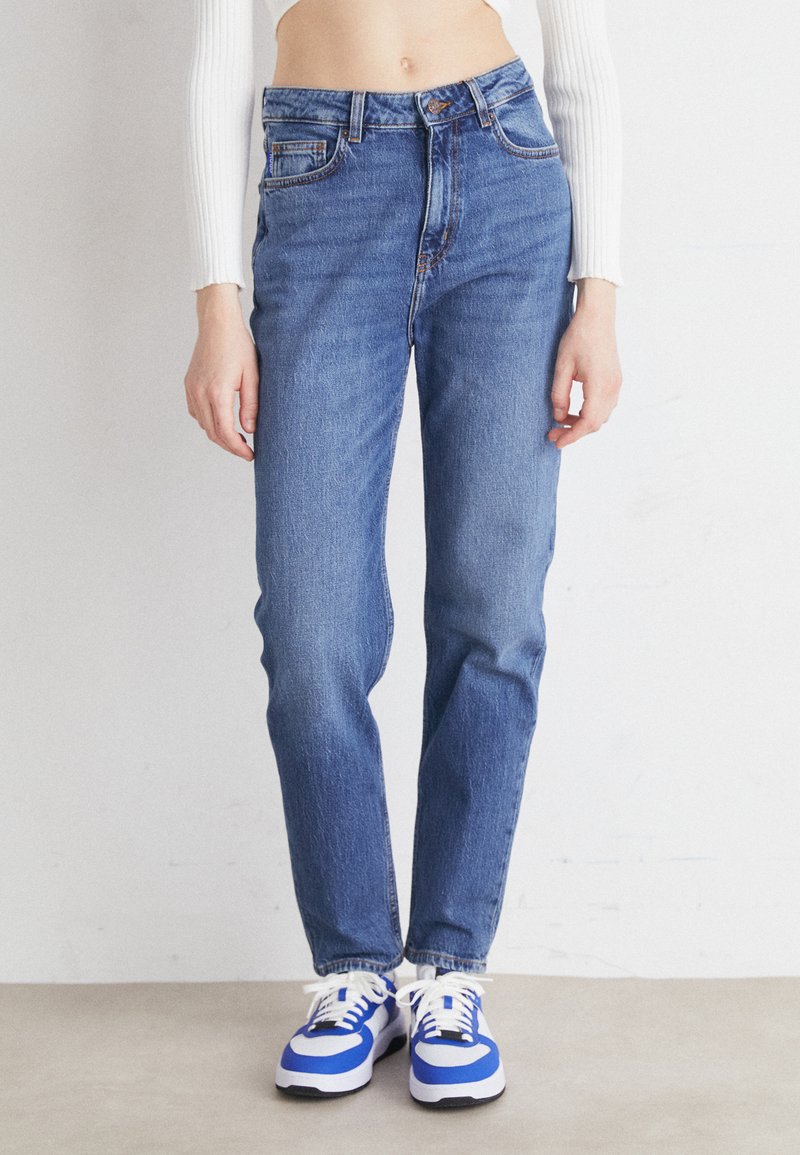 Jeans de mezclilla azul de cintura alta con pierna recta, diseño de cinco bolsillos y costuras en contraste, combinados con zapatillas azul y blancas.