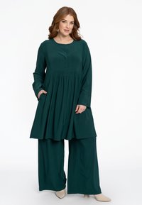 Türkise, langärmelige Tunika mit rundem Ausschnitt, Knopfdetails und plissierter Taille, kombiniert mit weitem Hosen. Weicher Stoff und sichtbare Taschen.