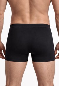 Vista posteriore di un uomo che indossa boxer aderenti neri semplici su uno sfondo bianco uniforme.