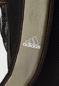 Sac à dos noir avec tissu en maille, un panneau texturé vert olive, logo Adidas blanc et accents de couture orange.
