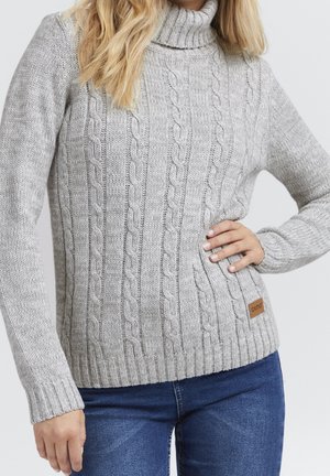 Femme portant un pull à col roulé torsadé gris clair et un jean bleu, main posée sur la hanche, sur un fond clair uni.